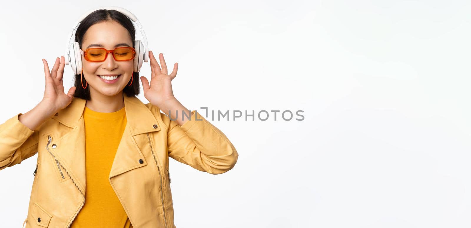 Beautiful asian girl, laughing happy, listening music in headphones, standing over white background. Copy space
