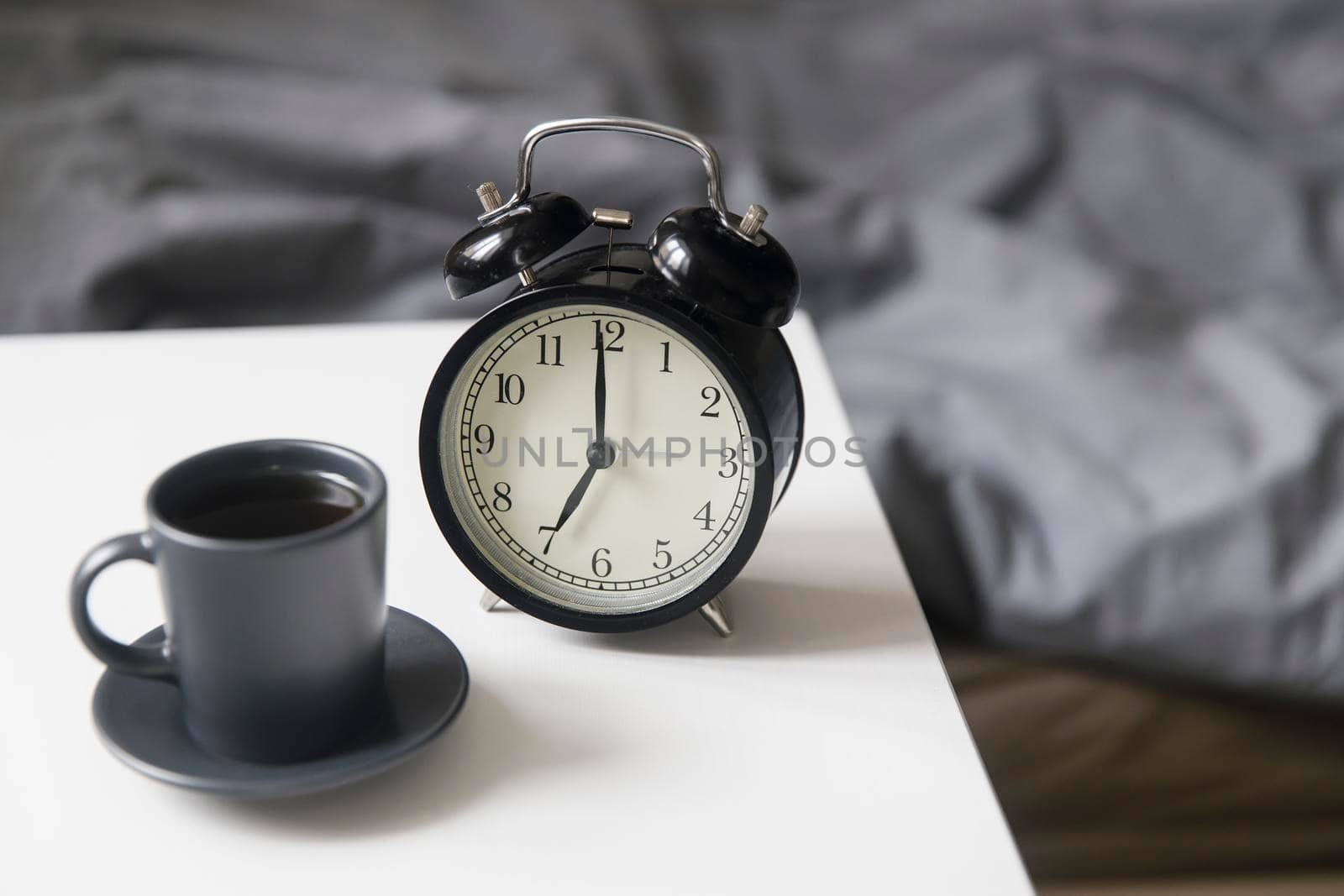 Image alarm clock with a black cup of coffee on a white bedside table in front of the bed with gray linens. The room is in beige tones.