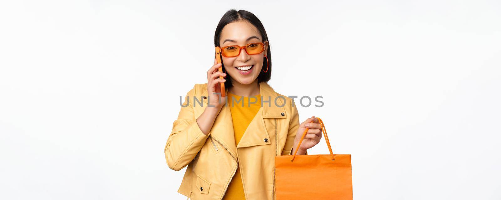 Modern korean girl on shopping, holding store bag, talking on mobile phone and smiling, standing over white background.