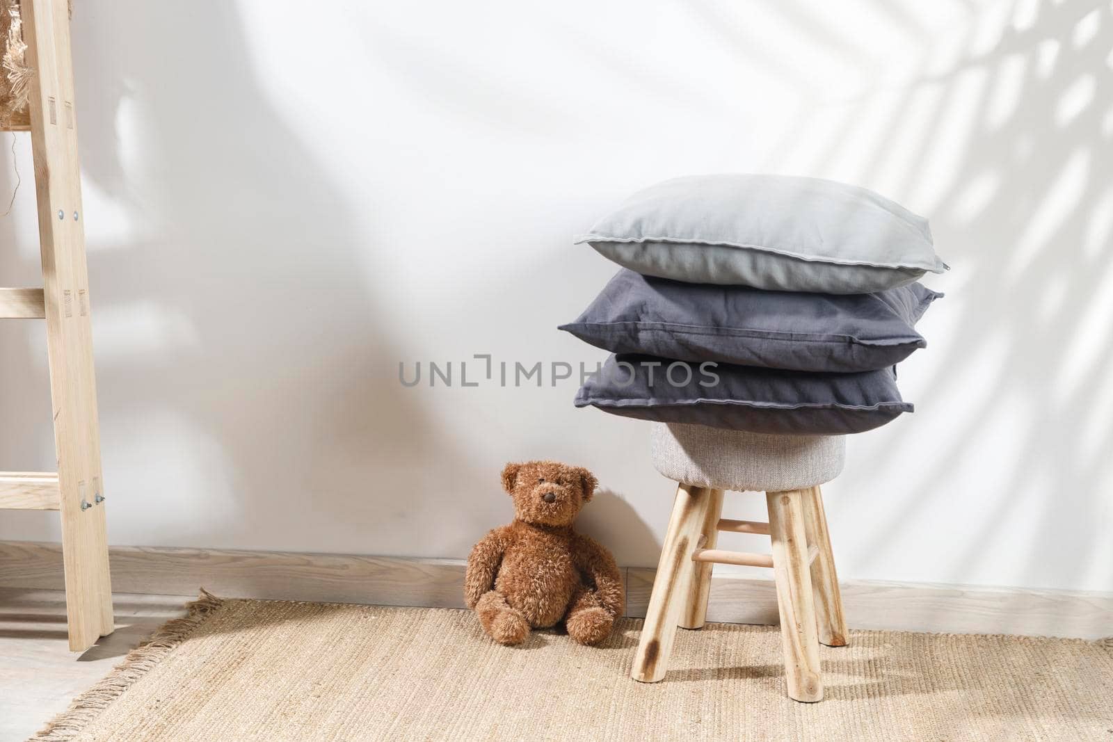 The teddy bear is leaning against the wall next to a small children's stool with three gray pillows on it. The stepladder is leaning against the wall. Copy space