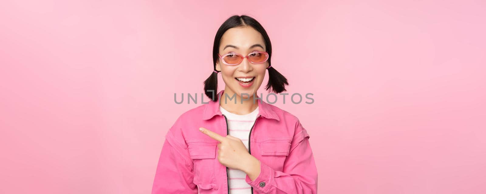Portrait of smiling asian girl in stylish outfit, sunglasses, pointing finger left, showing advertisement, banner, standing over pink background. Copy space