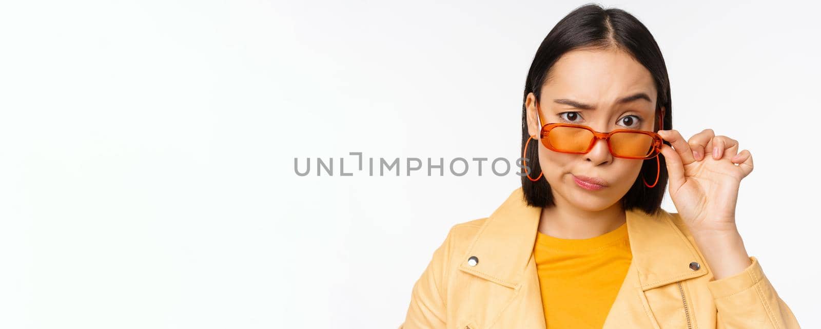 Image of young asian stylish woman, looking from under sunglasses with skeptical, disbelief face, express doubt, standing over white background. Copy space