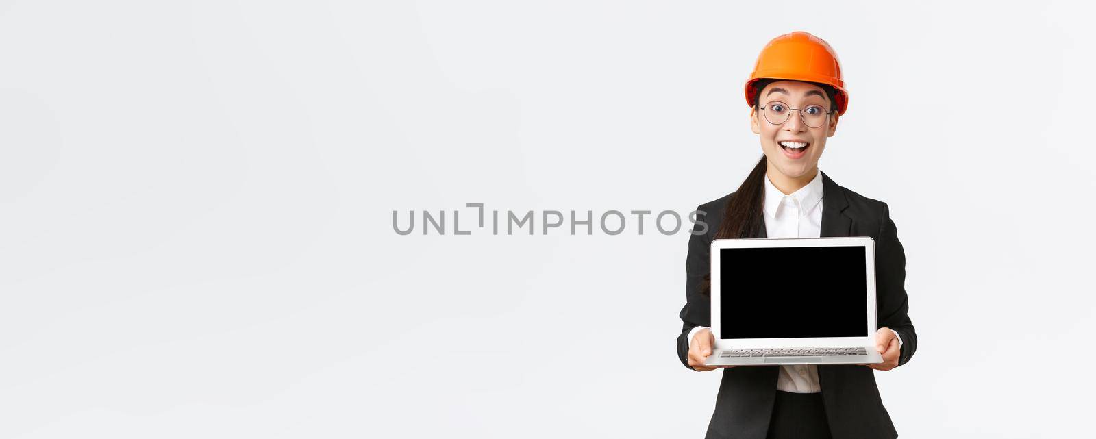 Excited happy asian female engineer, industrial manager in business suit and safety helmet showing laptop screen to investors or clients, amazed with great profit chart, introduce diagram.