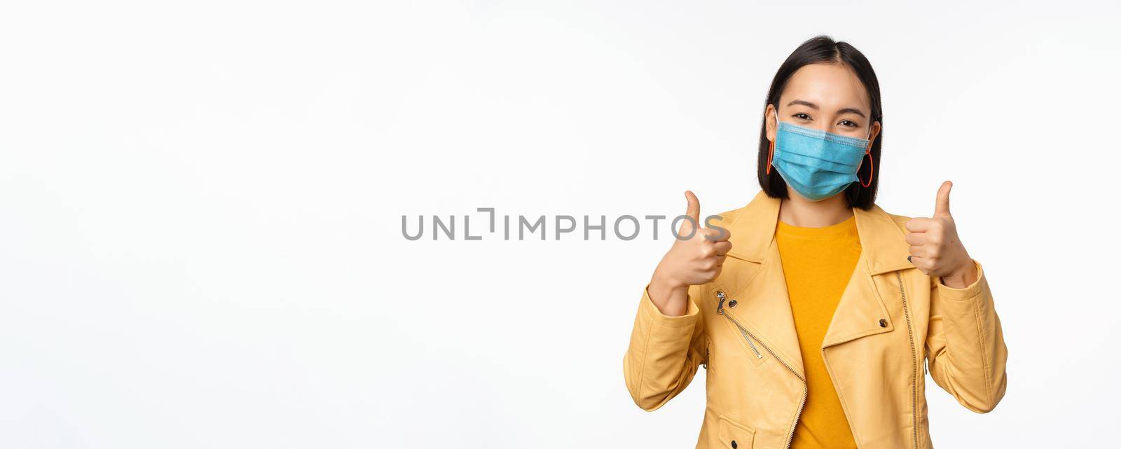 Smiling asian woman in medical face mask showing thumbs up, support wearing protective equipment from covid-19, coronavirus pandemic, white background.