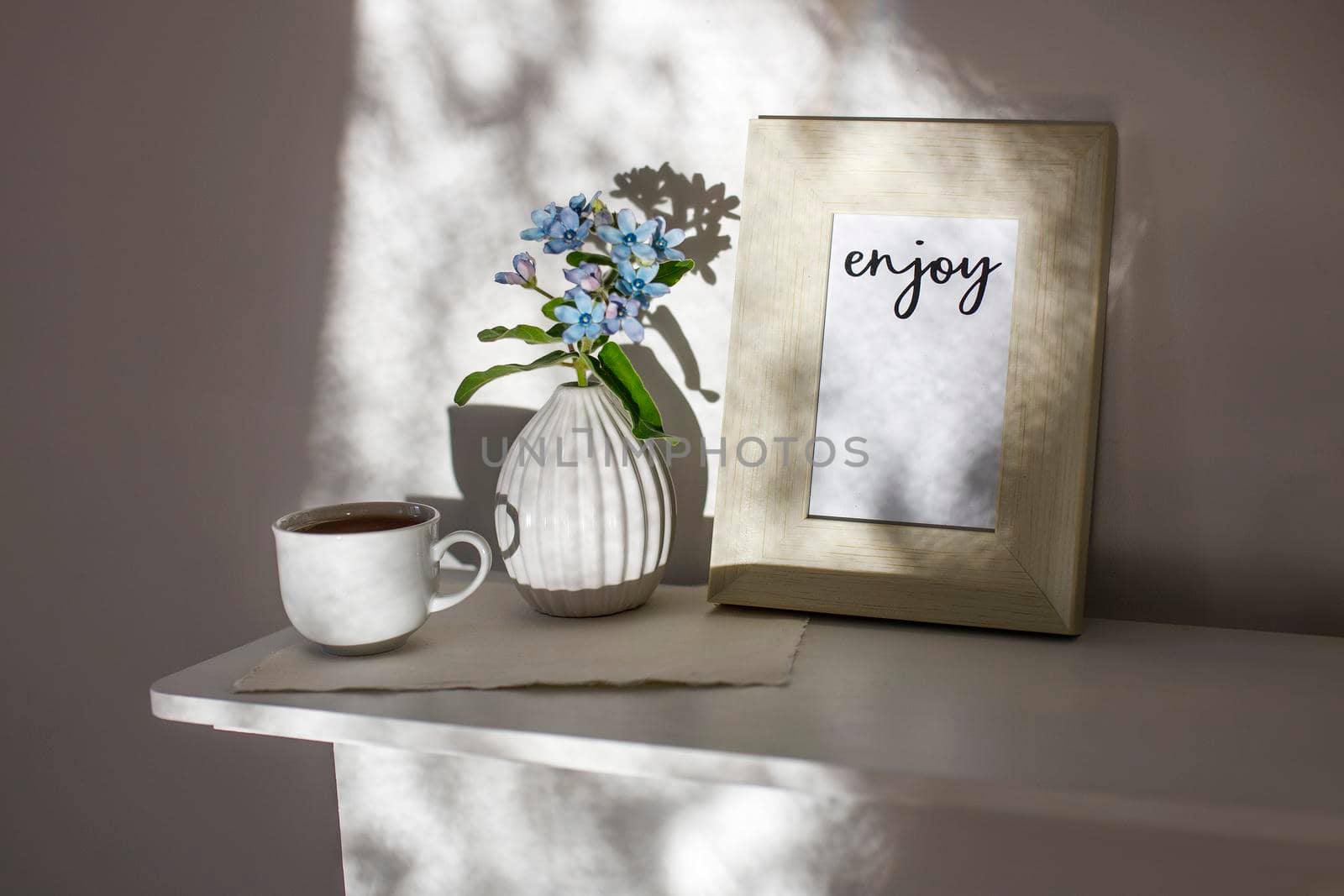 Flower hackelia velutina in a white fluted vase in the style of the seventies, a photo frame with the words Enjoy, and a cup of coffee on the dresser. Scandinavian style