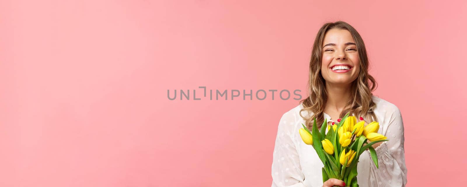 Spring, happiness and celebration concept. Close-up of happy and carefree blond european girl receive beautiful bouquet of flowers, holding yellow tulips, smiling and laughing, pink background.