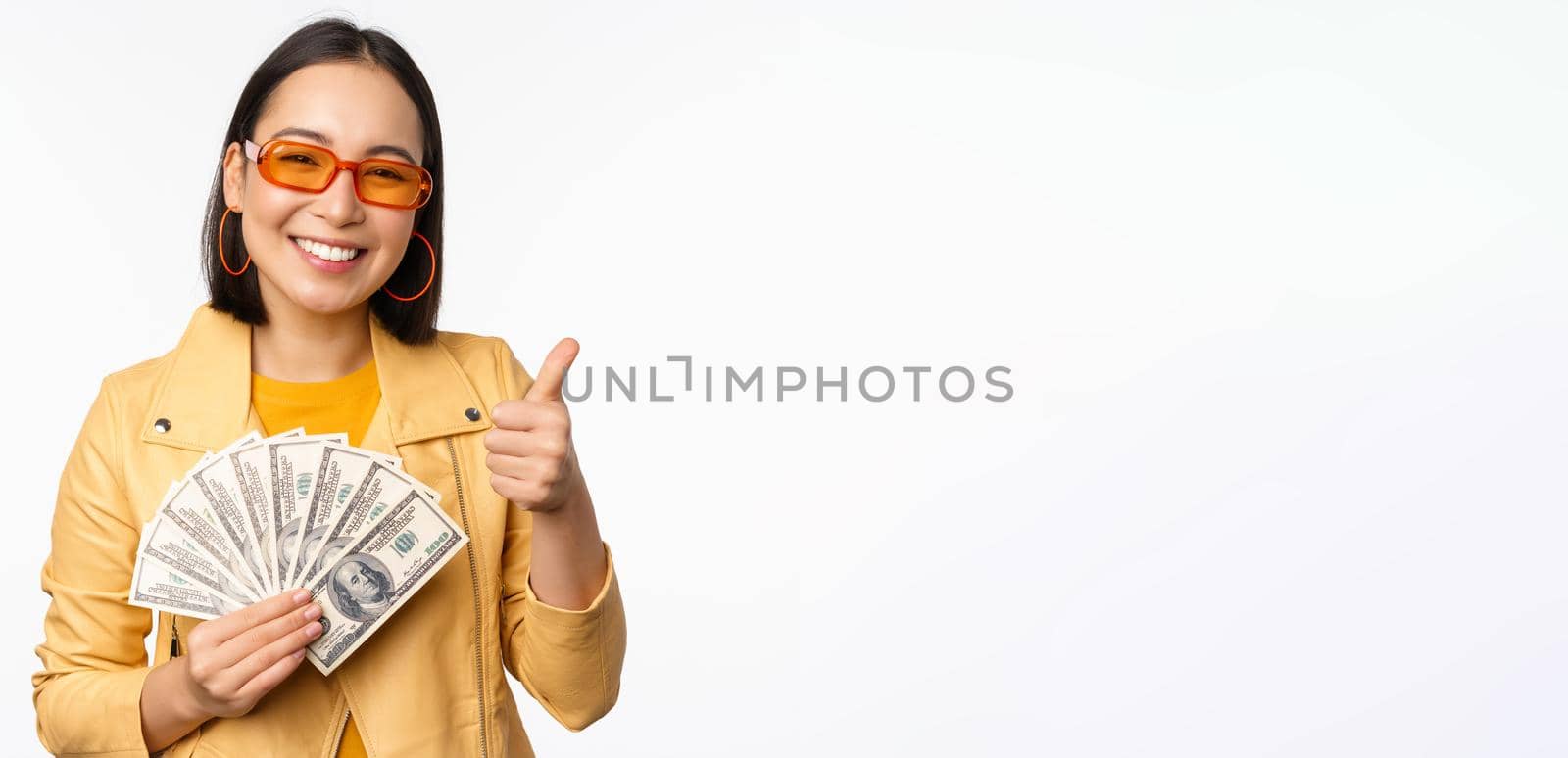 Stylish smiling asian girl holding money cash, showing dollars and celebrating, standing over white background. Copy space