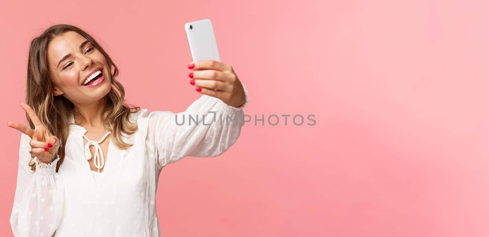 Close-up portrait of cheerful lovely, feminine blond girl in white dress, taking selfie on mobile phone, make kawaii peace sign while take photo, capturing spring moment, pink background.