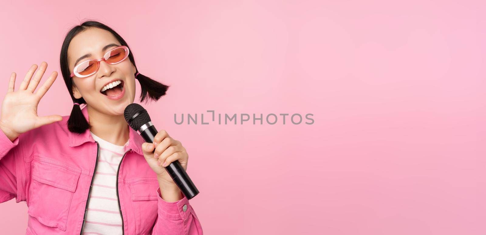 Happy beautiful asian girl singing with mic, using microphone, enjoying karaoke, posing against pink studio background.