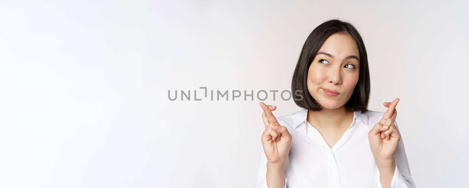 Hopeful young lucky girl making wish, cross fingers and looking aside with thinking face expression, white background.