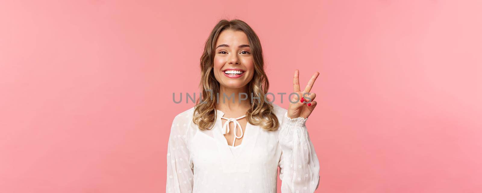 Close-up portrait of romantic lovely smiling girl with blond short hair, wearing white dress, show peace sign enjoying spring, grinning and look camera over pink background. Copy space