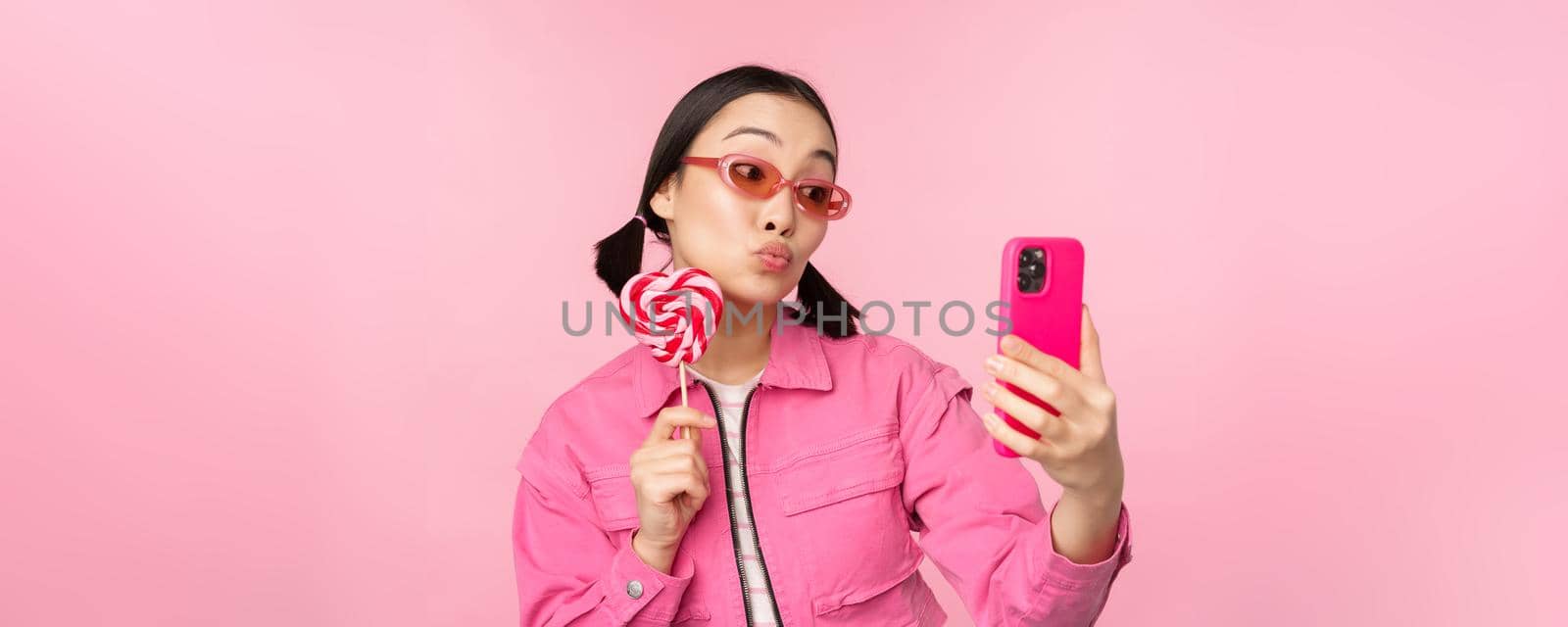 Portrait of stylish, happy asian girl taking selfie with candy, lolipop sweets and smiling, taking photo with mobile app, standing over pink background.