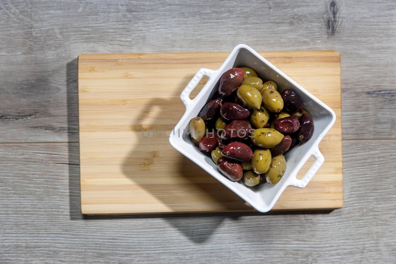 Deep square porcelain bowl with green and dark olives on a wooden board with shadow from a palm tree on a blue background