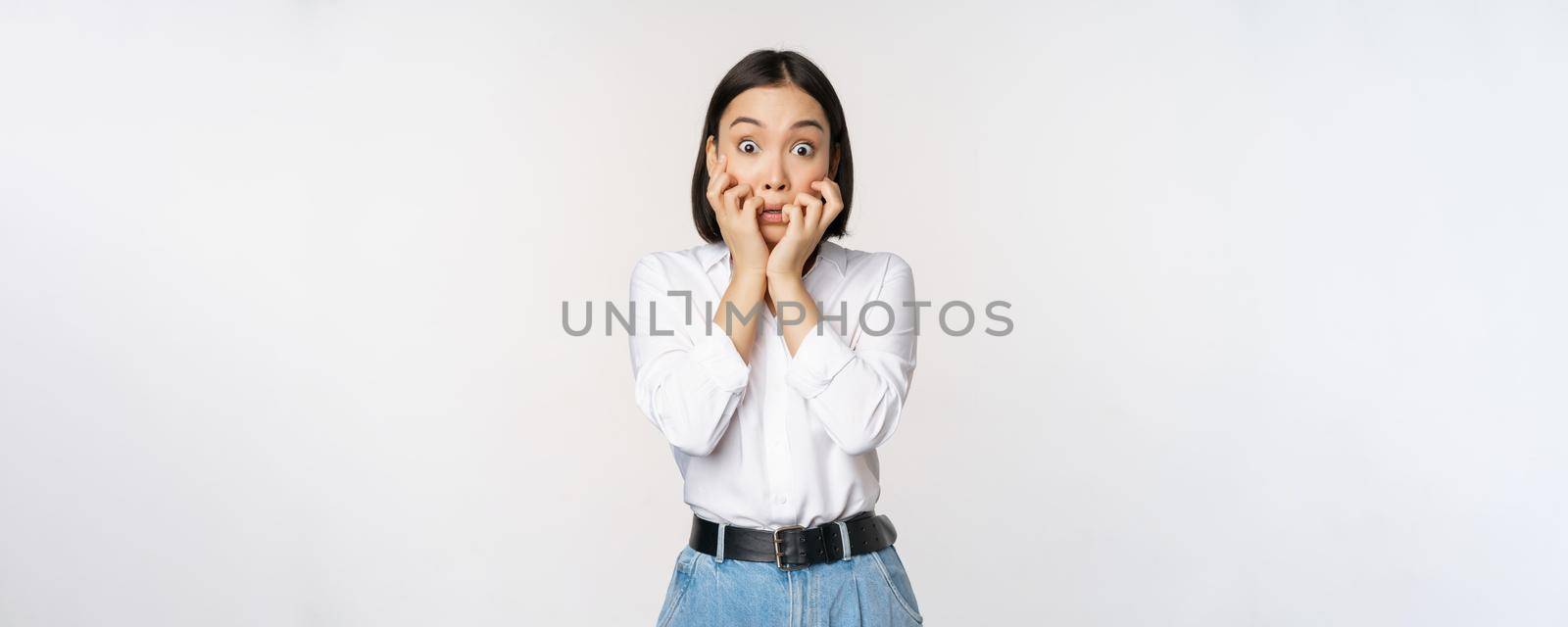 Portrait of scared asian woman watching smth scary, biting fingers on hands and looking at camera frightened, standing over white background.