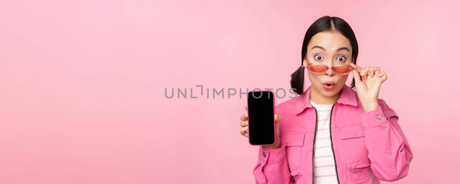 Portrait of asian girl showing mobile phone screen, reacting surprised, standing over pink background. Copy space