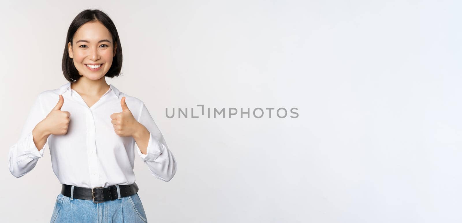 Image of beautiful adult asian woman showing thumbs up, wearing formal office, university clothing, recommending company, standing over white background.