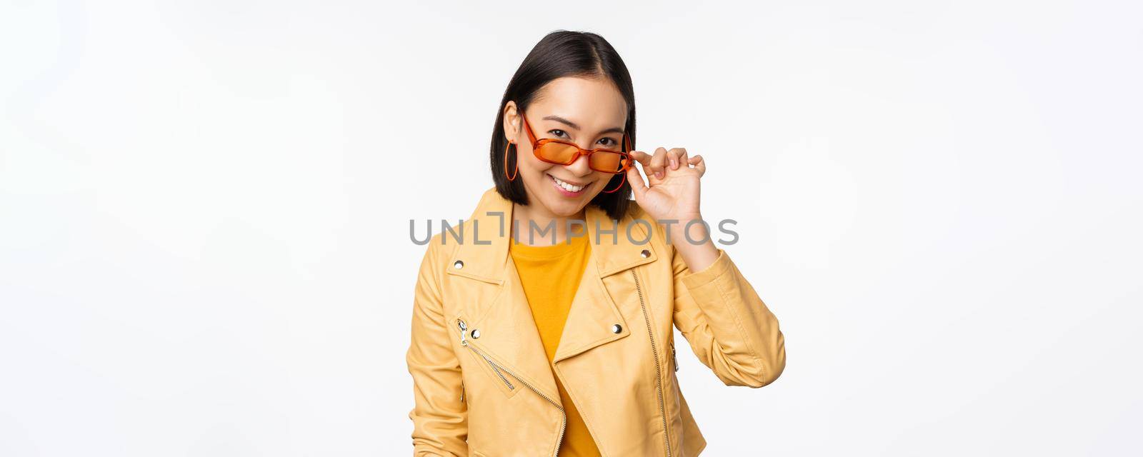 Beautiful asian girl in sunglasses, smiling and looking happy, posing in yellow jacket against white background. Copy space