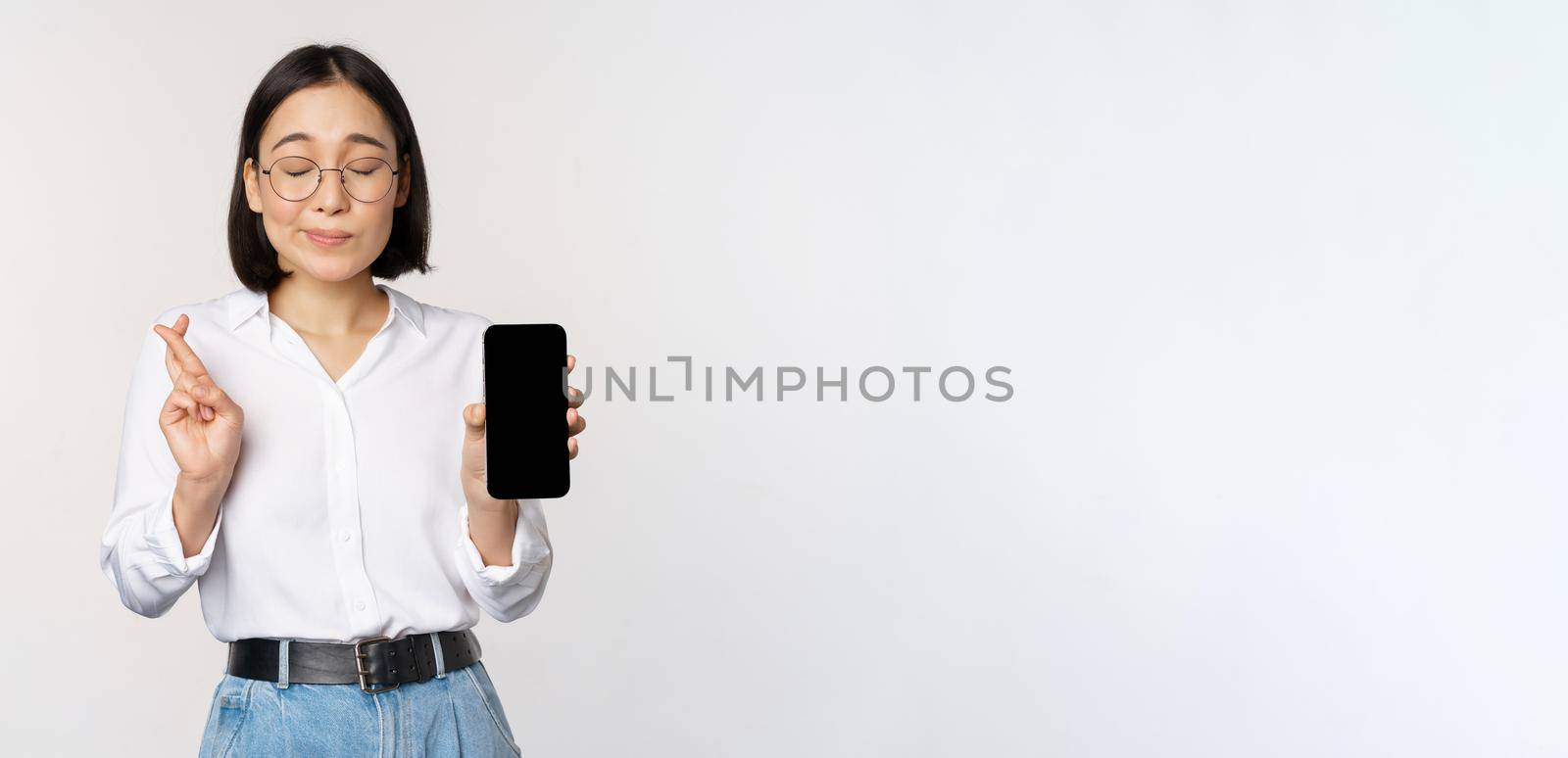 Hopeful young asian woman, showing mobile phone screen, app interface and fingers crossed, hoping for smth, making wish or paying, standing over white background by Benzoix