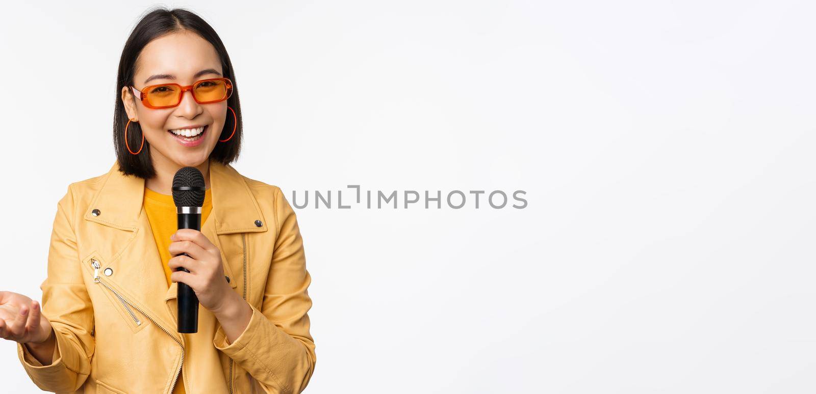 Portrait of beautiful asian woman in sunglasses, stylish girl singing, giving speech with microphone, holding mic and smiling, standing over white background. Copy space