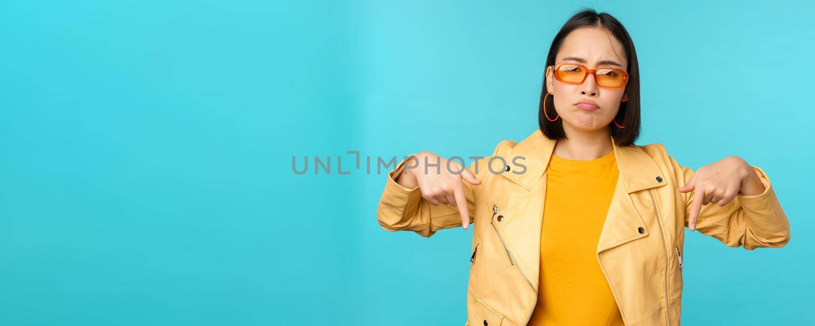Sad and gloomy chinese girl in sunglasses, points fingers down and grimaces, looks miserable and disappointed, emotion of regret, standing over blue background.