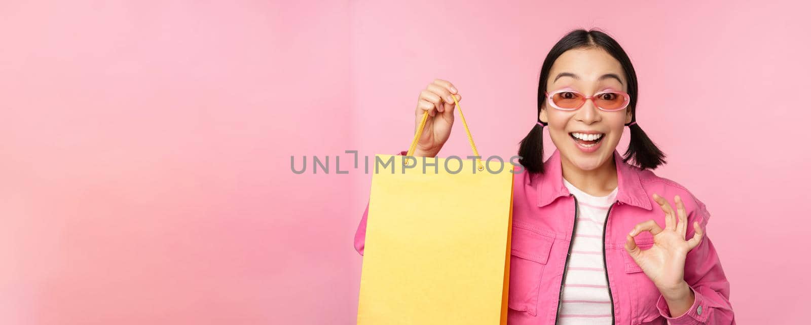 Shopping. Stylish asian girl in sunglasses, showing bag from shop and smiling, recommending sale promo in store, standing over pink background.