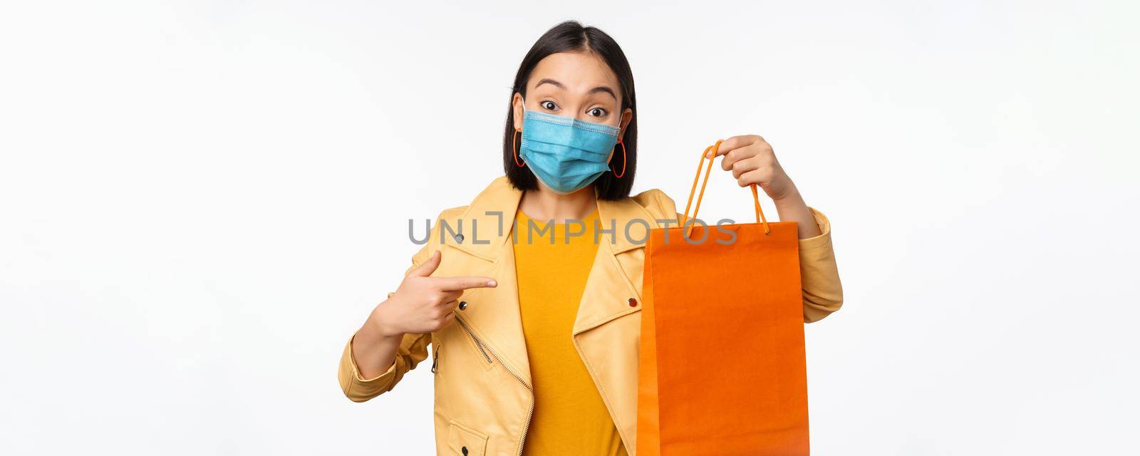 Covid-19 and shop concept. Young asian stylish woman, wearing medical face mask, holding shopping bag, going in malls during pandemic, white background.