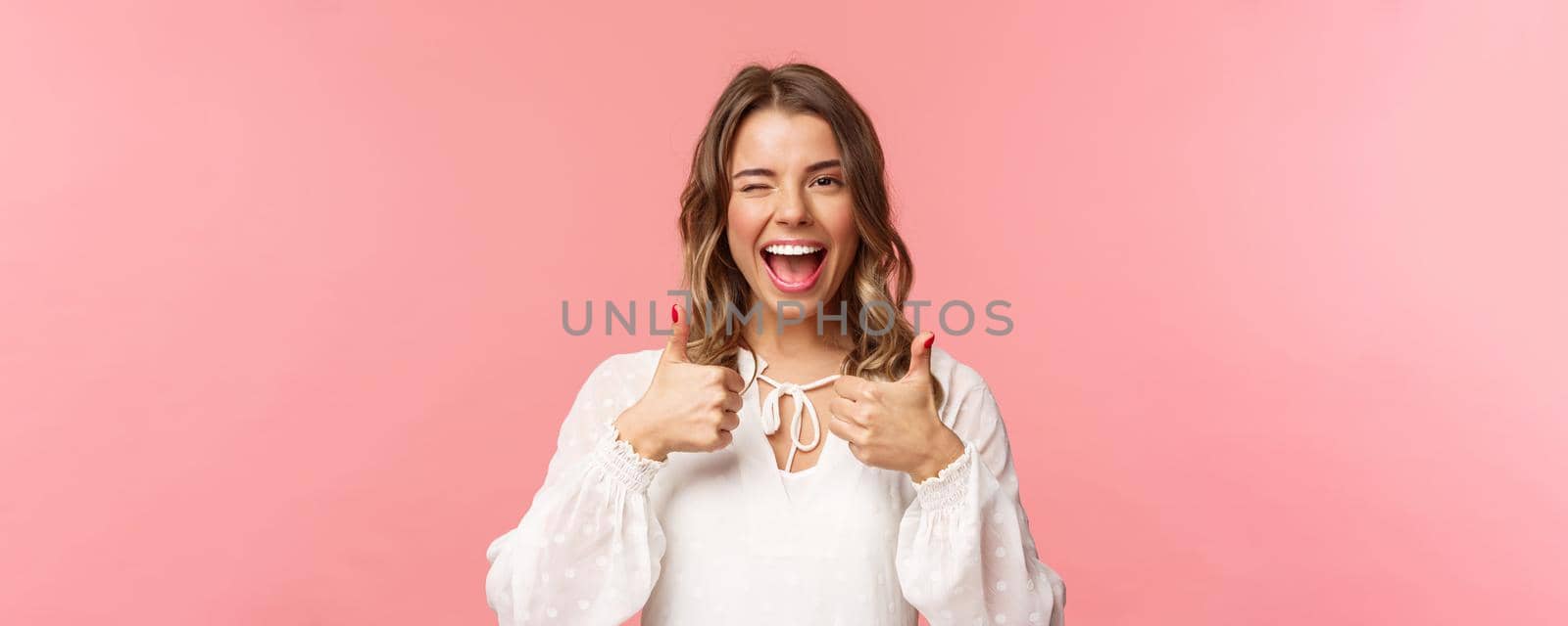 Portrait of sassy good-looking blond girl assure all good, show thumbs-up and wink with classy pleased smile, recommend visit place, shop at online store, standing in white dress pink background.