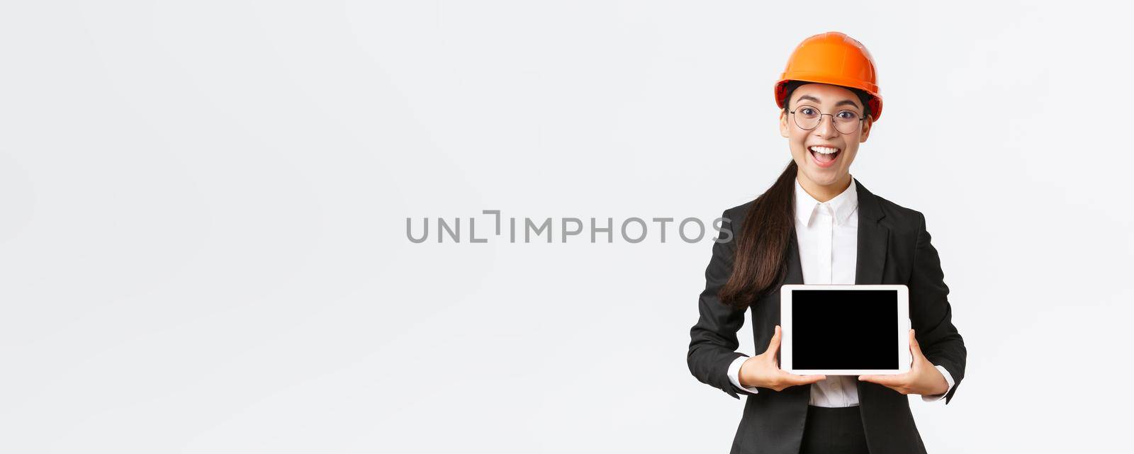 Excited and amazed smiling asian female construction engineer, architect in safety helmet and business suit, showing digital tablet screen with amazed expression, standing white background.