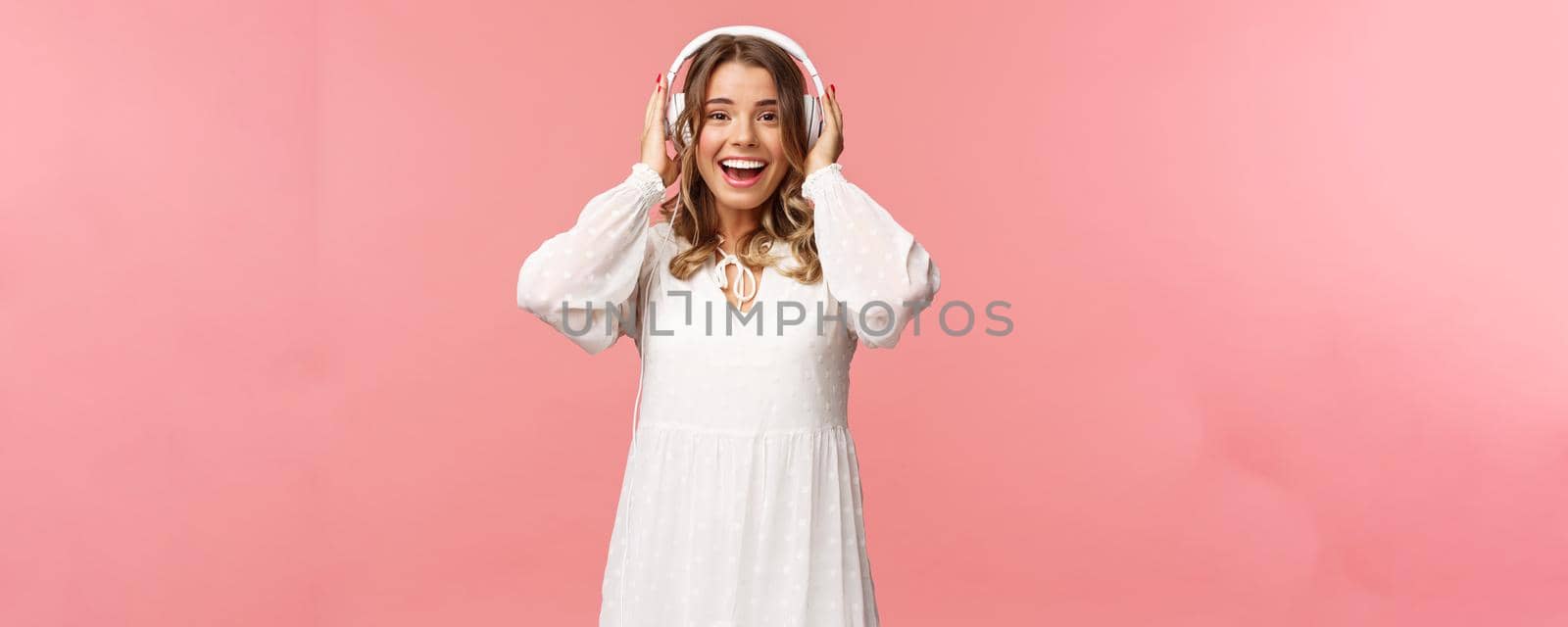 Portrait of excited, happy good-looking girl in white tender dress, wearing headphones and smiling amazed as looking at camera, fascinated with good sound quality, pink background.