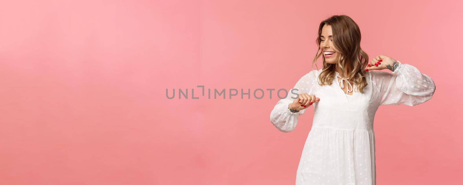 Beauty, fashion and women concept. Tender and carefree pretty young girl enjoying spring time, wearing white dress, dancing and looking away with beaming smile having fun, pink background.