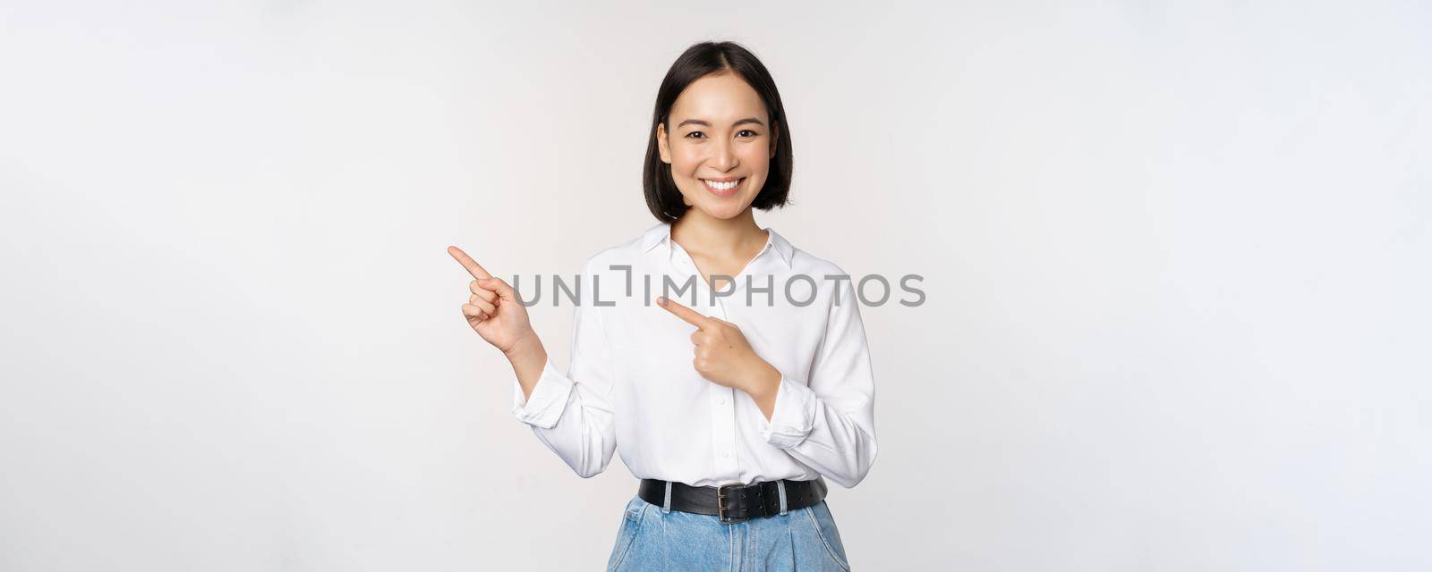 Image of smiling young office lady, asian business entrepreneur pointing fingers left, showing client info, chart of banner aside on copy space, white background by Benzoix