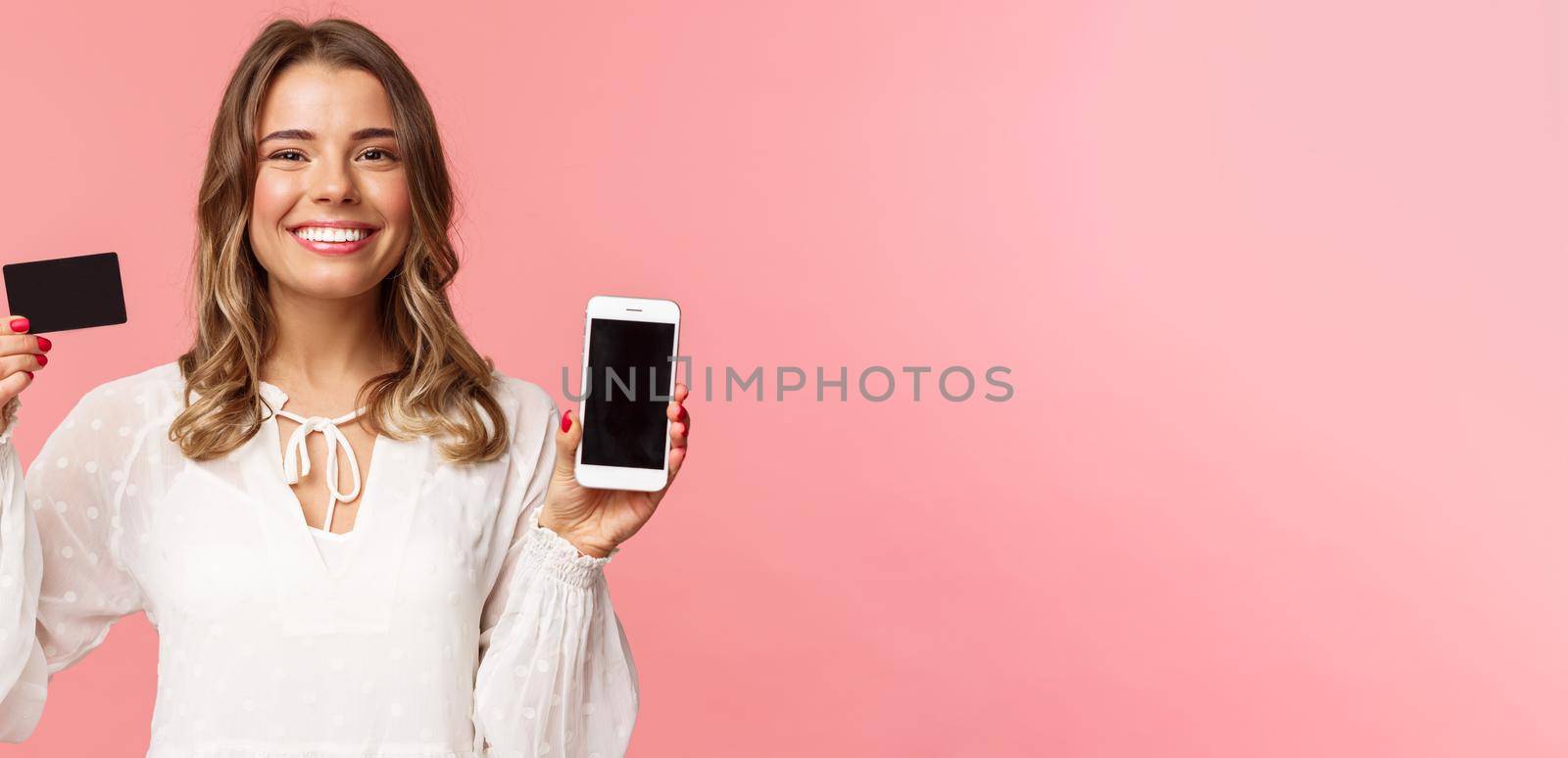 Finance, shopping and technology concept. Close-up portrait of cheerful, romantic blond cute girl in white dress, holding credit card and mobile phone, showing smartphone display, application promo.