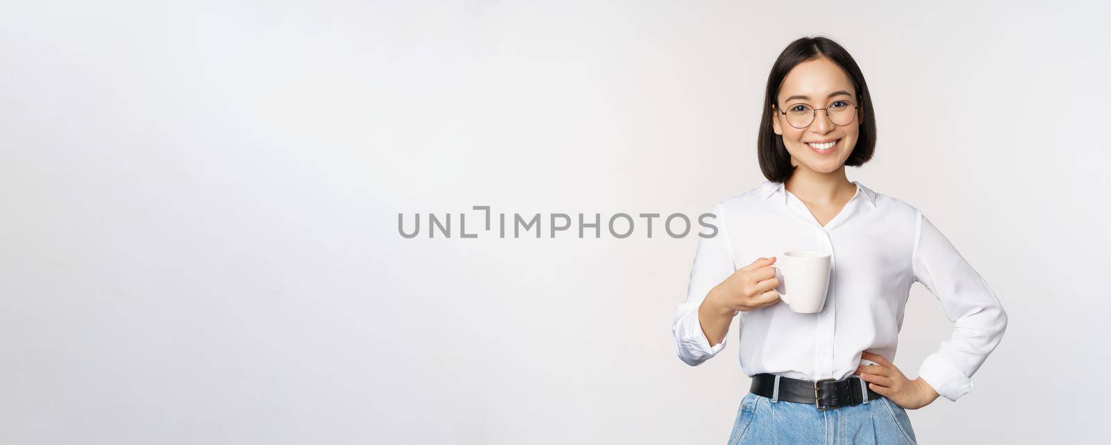 Happy young energetic asian woman smiling, drinking, holding cup mug of coffee, standing confident against white background.