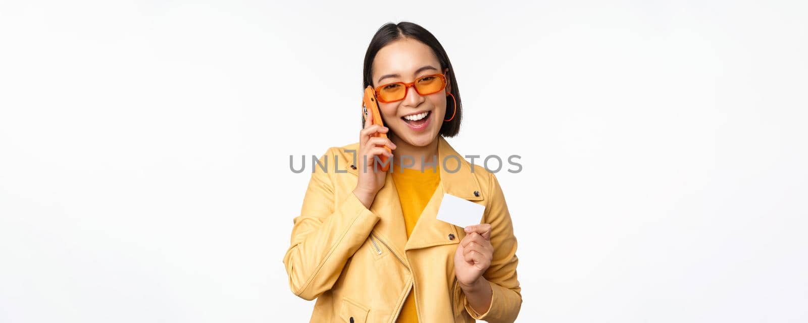 Stylish asian female model, talking on smartphone and showing credit card, standing over white background. Copy space
