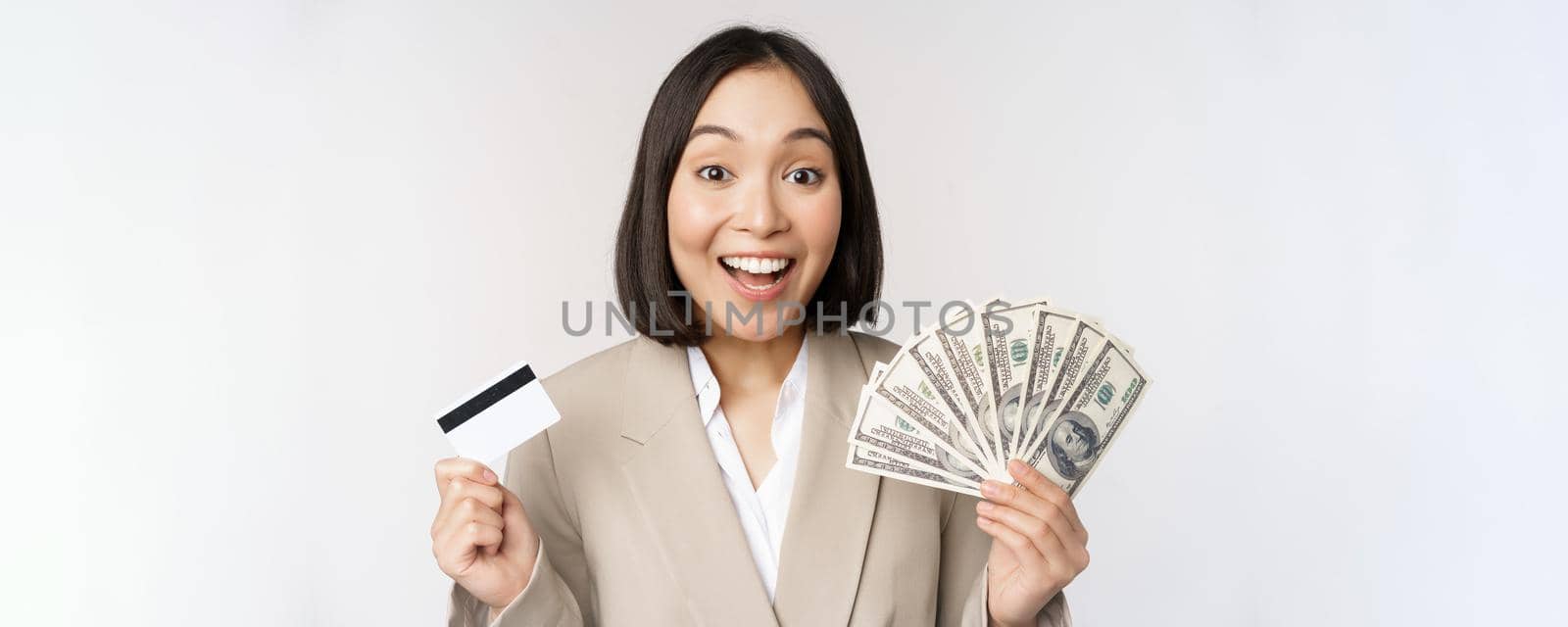 Enthusiastic young asian businesswoman, showing money dollars and credit card, standing over white background.