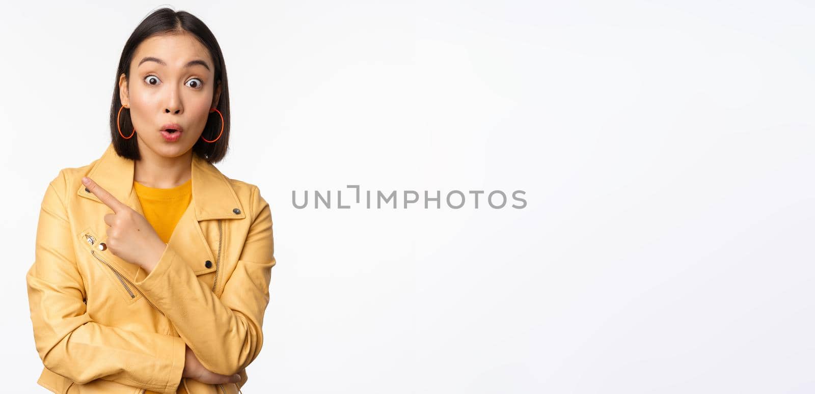 Portrait of beautiful asian woman express intereest and excitement about advertisement, store discount, pointing finger left, looking amazed, standing over white background.