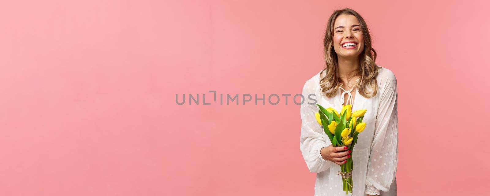 Holidays, beauty and spring concept. Carefree happy attractive blond girl in white dress holding yellow tulips and laughing with pleased expression, standing pink background, have romantic date by Benzoix