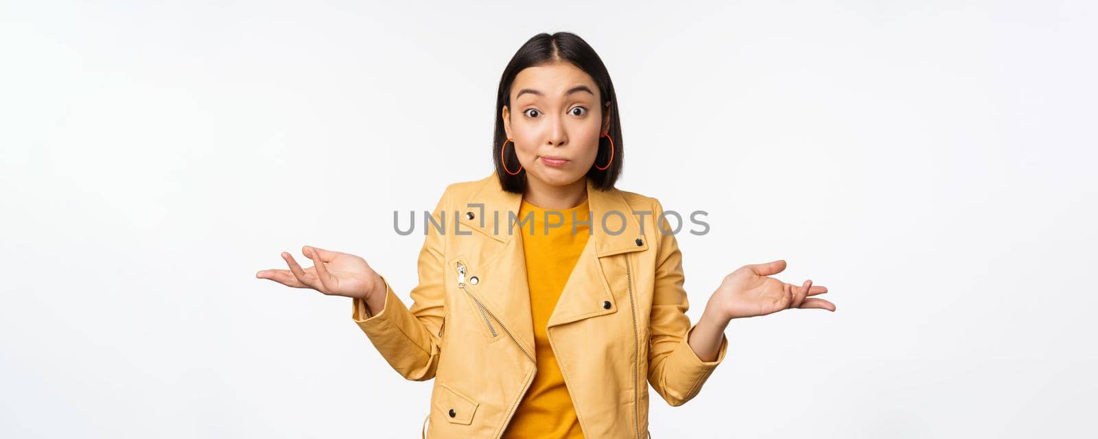 Image of korean girl shrugs shoulders and looks confused, dont know, cant tell, standing over puzzled over white background.