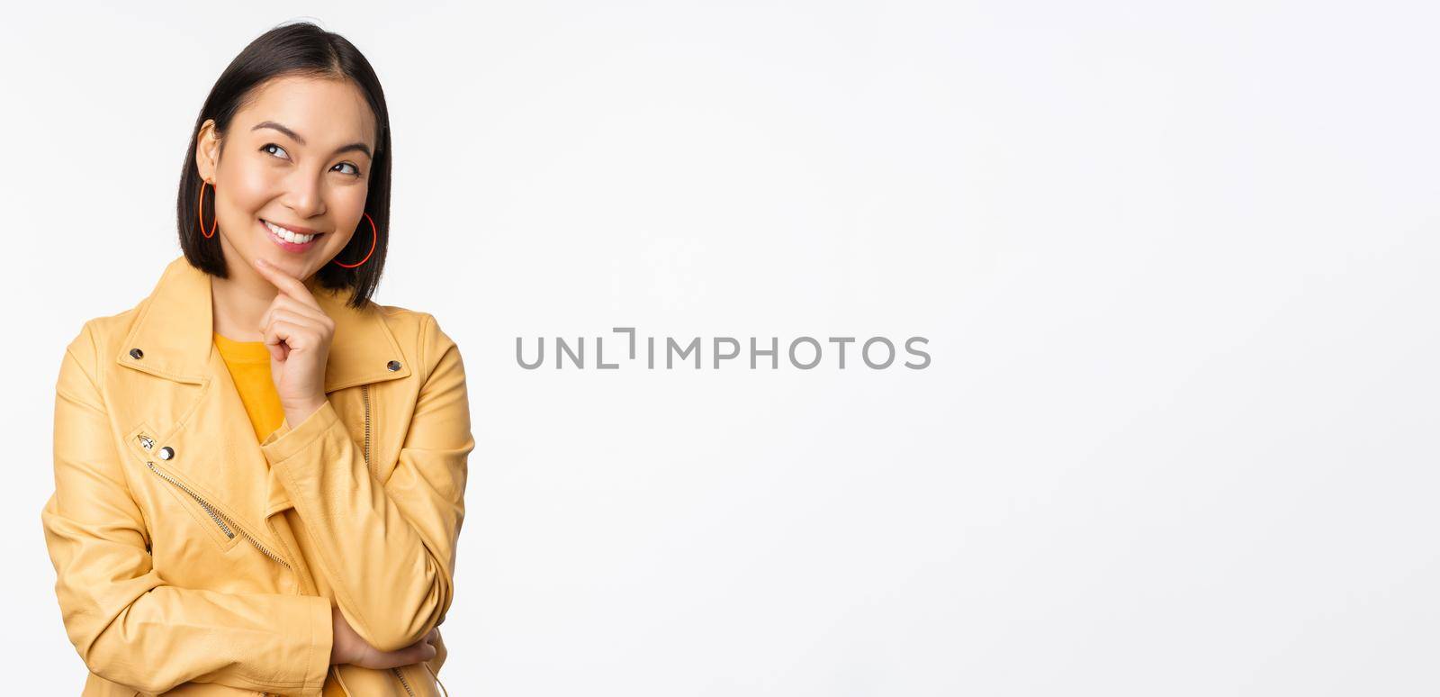 Portrait of stylish korean woman in yellow jacket, smiling thoughtful, thinking and looking up at logo or advertisement, standing over white background.