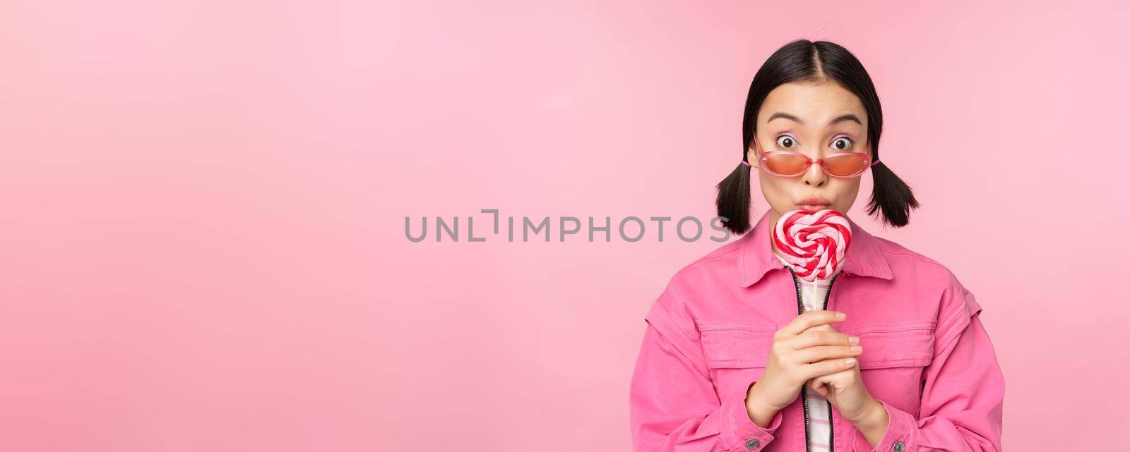 Silly and cute asian female model licking lolipop, eating candy sweet and smiling, looking excited, standing over pink background.
