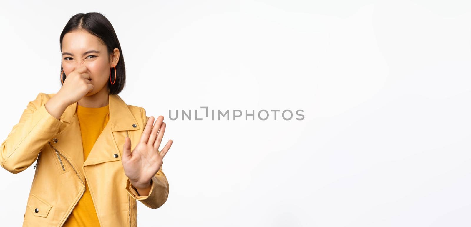 Image of asian girl shuts nose and reject smth with disgusting bad smell, shuts nose, standing over white background. Copy space