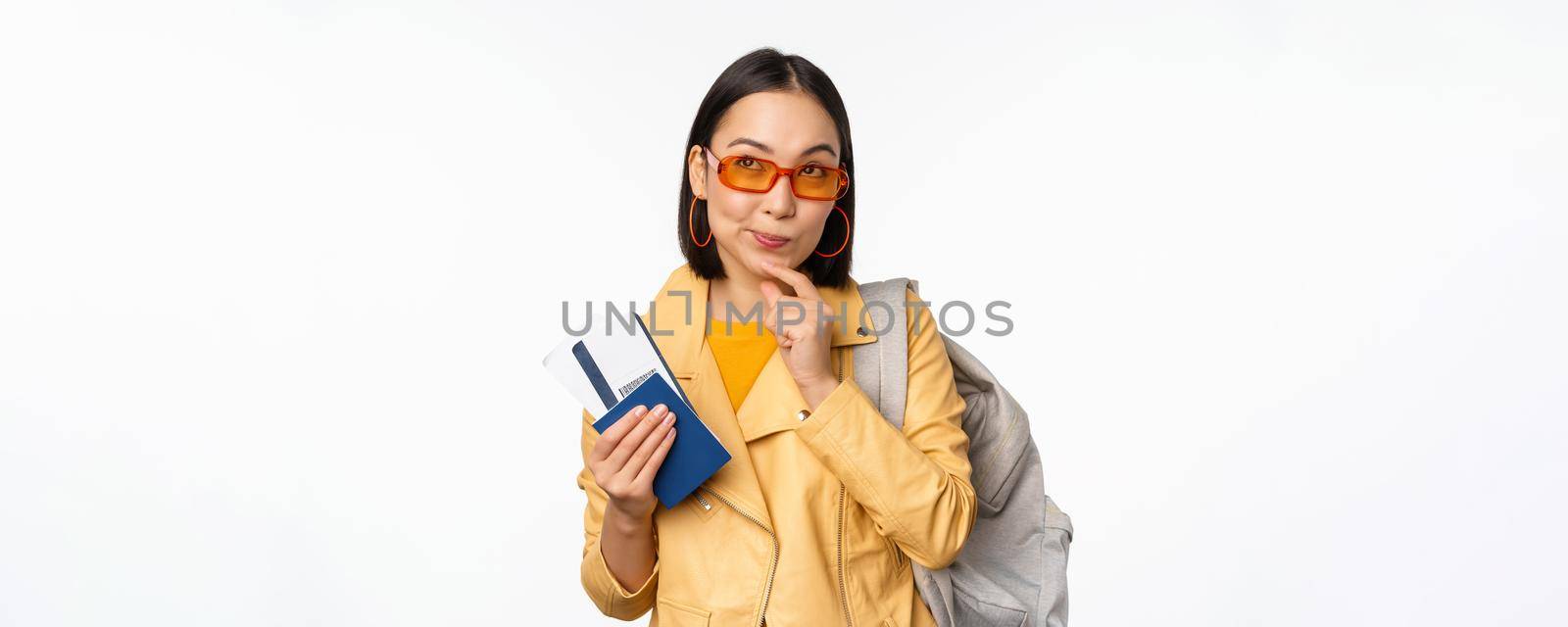 Asian girl tourist with boarding tickets and passport going abroad, holding backpack, thinking of travelling, standing over white background.