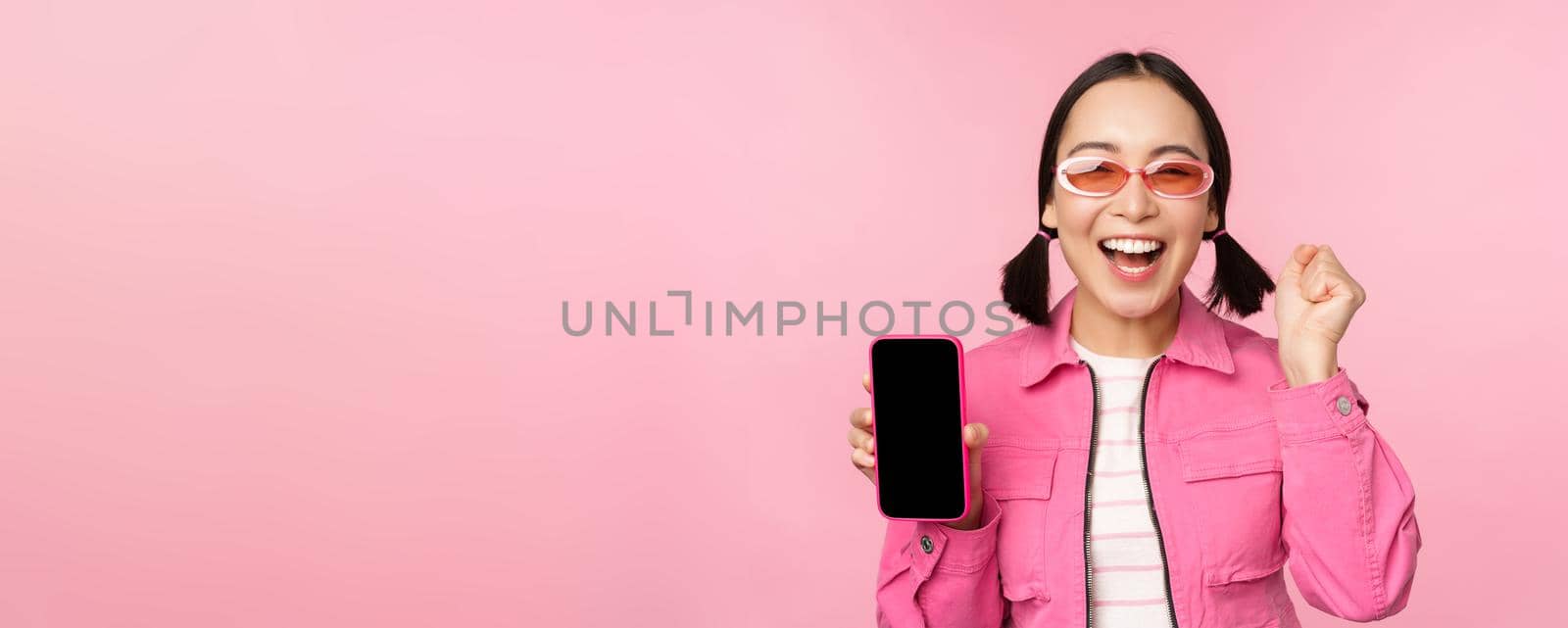 Excited asian girl laughs and smiles, shows mobile phone screen, smartphone application, standing over pink background.