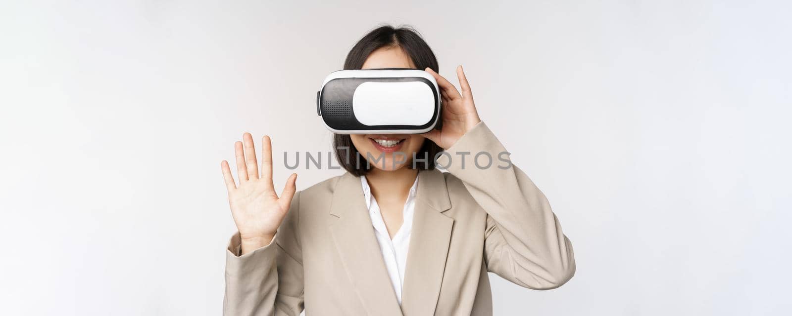 Meeting in vr chat. Asian businesswoman in virtual reality glasses, raising hand and saying hello, greeting someone, standing over white background.