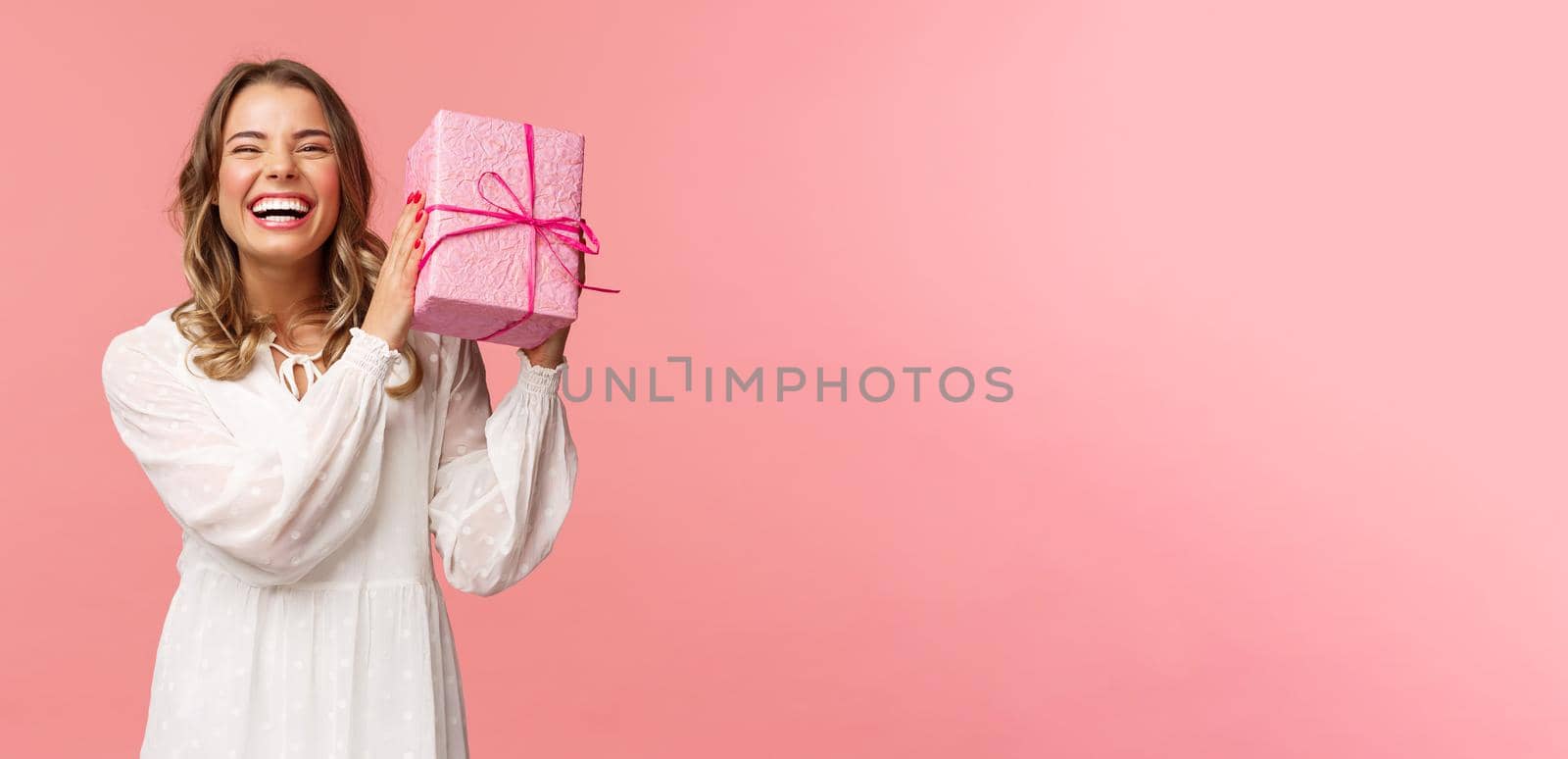 Holidays, celebration and women concept. Portrait of happy charismatic blond girl shaking gift box wondering whats inside as celebrating birthday, receive b-day presents, pink background.