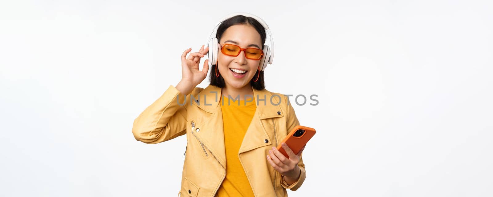 Happy stylish asian woman in sunglasses, listening music in headphones, holding smartphone, singing and dancing, standing over white background.
