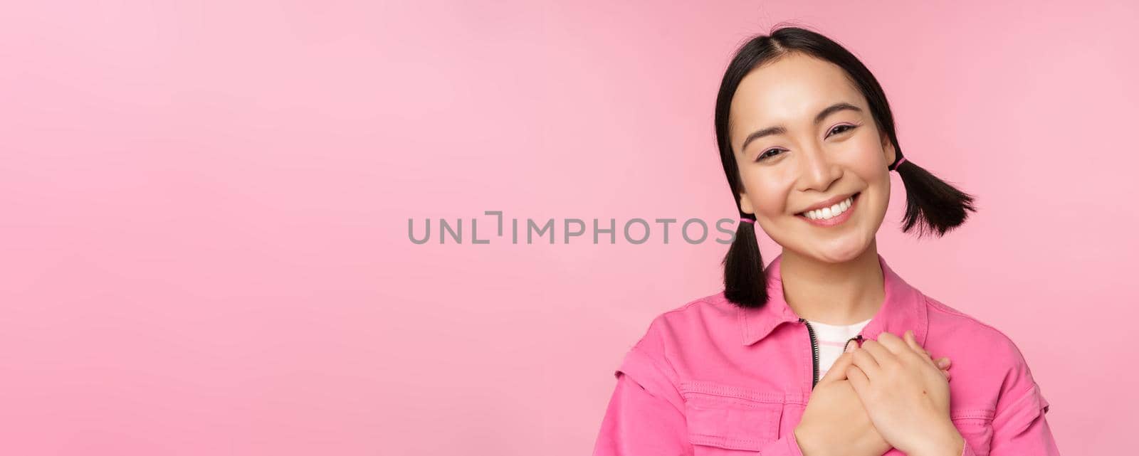 Cute korean girl with silly hairbuns, looking happy and grateful, thank you pose, holding hands on heart flattered, posing against pink background.