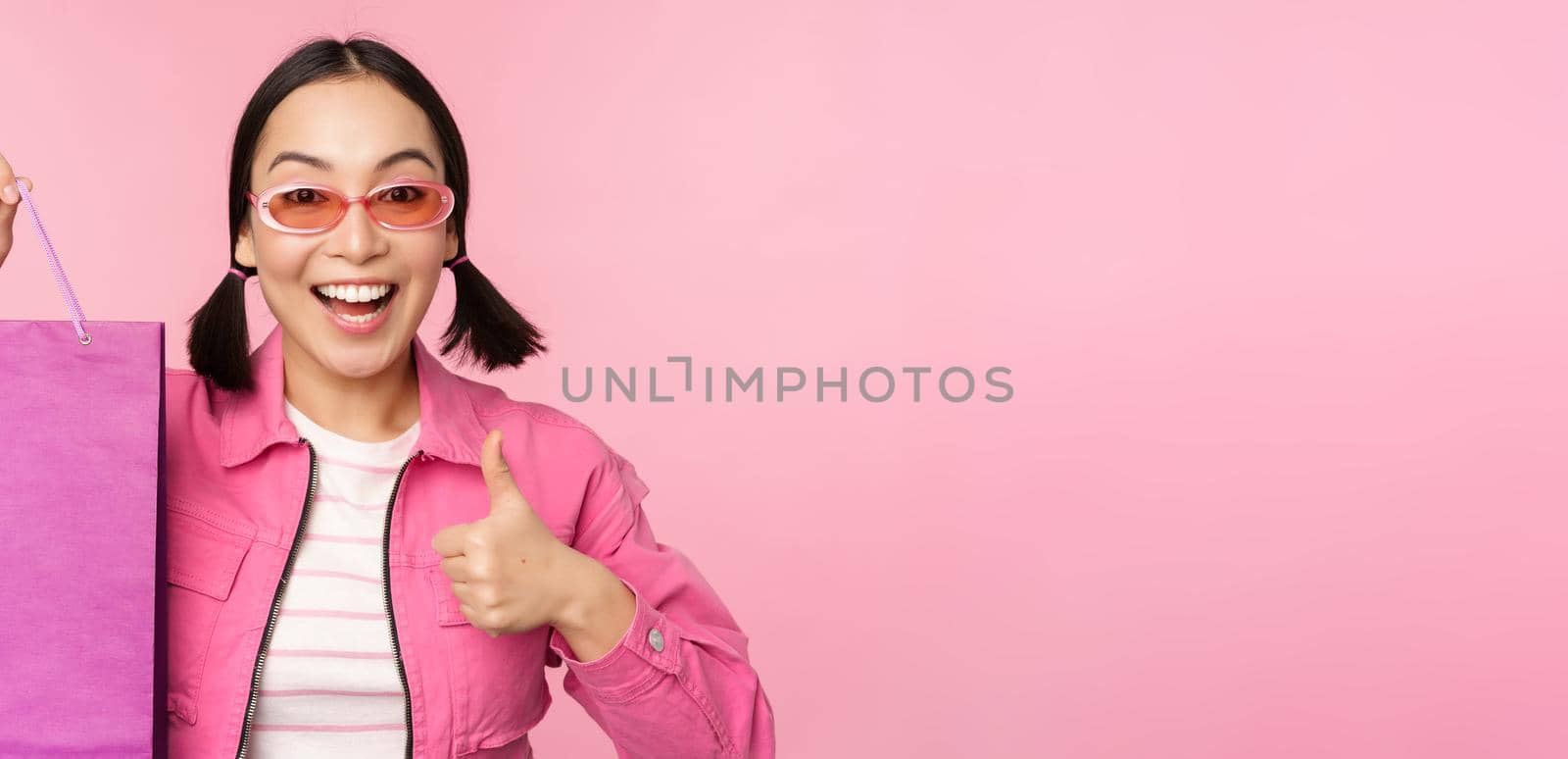 Shopping. Stylish asian girl in sunglasses, showing bag from shop and smiling, recommending sale promo in store, standing over pink background.
