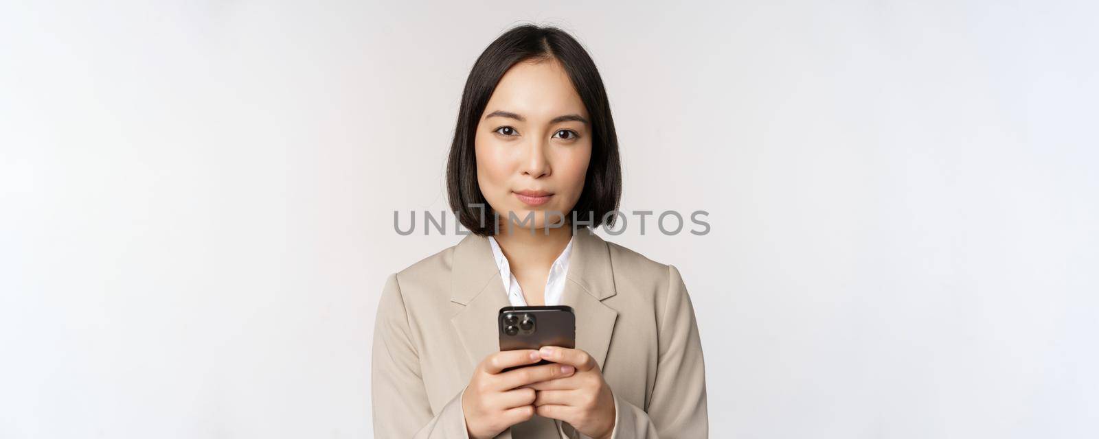 Image of asian businesswoman in suit, holding mobile phone, using smartphone app, smiling at camera, white background.