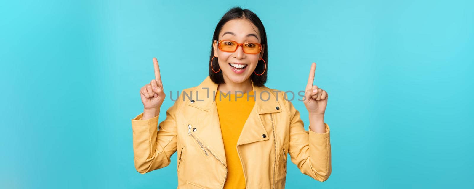 Enthusiastic asian girl in sunglasses, points fingers up, shows banner or logo on top, stands over blue background. Copy space